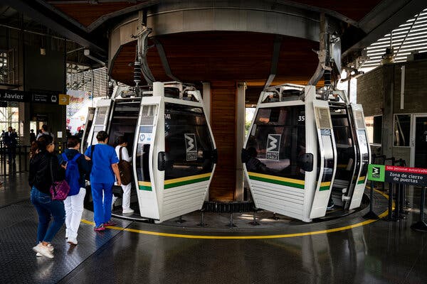  Two cable cars parked on the ground. Four people line up to enter one of the cable cars, which are white with two yellow and green stripes. The cable cars are connected to a gray structure. 