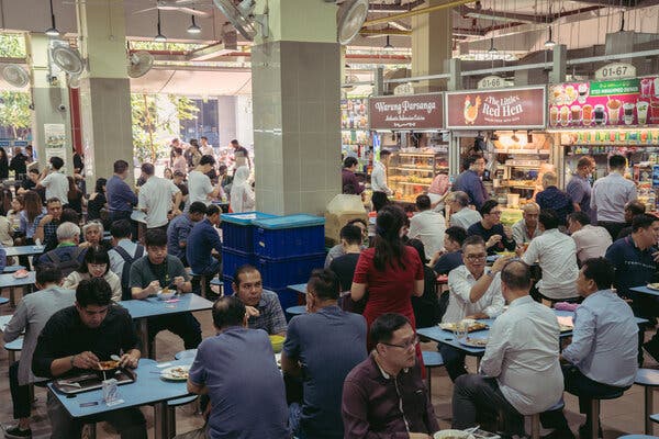 Several people sitting and eating together at communal blue tables with blue stools. Behind them are food stalls. 