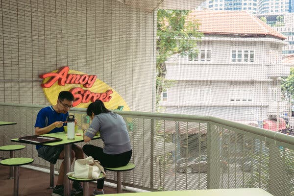 A man and a woman eat together on a balcony, while sitting at a bright green table with green stools. Behind the balcony is a sign that reads “Amoy Street.” 